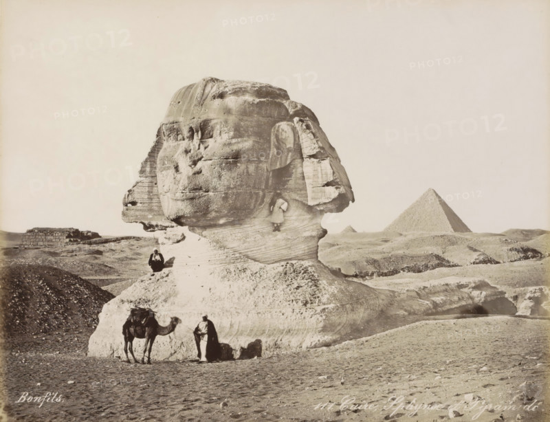 Sphinx and pyramids of Giza, Caire, Sphynx et Pyramide, photograph, Part of Travel album with 80 photos of Egypt., negative number: '111', photographer: Maison Bonfils, Gizeh, c. 1870 - c. 1898, North Africa, Giza, pasteboard, paper, albumen print, height 220 mm x width 282 mm, pyramid (historical grave form), piece of sculpture, reproduction of a piece of sculpture (sculpture), A large weathered stone sculpture of a human head and neck dominates the foreground, showing layered textures and erosion lines on its surface. A person sits on the stone near the sculpture’s base, and another person stands in front of the base beside a camel, which has a saddle and some coverings. A small figure is positioned on a ledge on the sculpture’s neck. Low rocky hills and sandy ground surround the sculpture, and a pyramid with a smooth sloped face rises in the distant background against a pale sky.