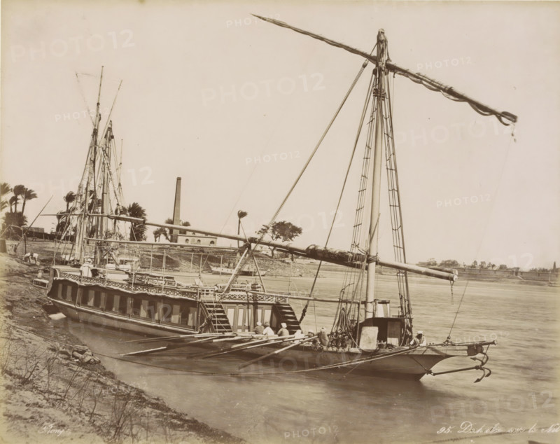 Dahabie on the Nile, Passenger boat (dahabie) on the Nile, photograph, Part of Travel Album with 80 Photos of Egypt., negative number: '95', photographer: Maison Bonfils, Egypt, c. 1870 - c. 1898, North Africa, pasteboard, paper, albumen print, height 222 mm x width 278 mm, sailing-ship, sailing-boat, Nile, A large boat with tall masts and long spars is docked along a riverbank, showing a wooden hull with a row of cabin windows and an open deck with railings and steps leading down; several ropes, rigging lines, and ladders run up the masts, and a group of people wearing light clothing and head coverings sit or stand near the stern and along the deck; the river water is calm and stretches across the image with another boat partly visible in the background; palm trees and a building with a tall chimney stand on the far shore, and the sky above is clear and pale.