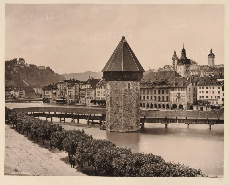 View of the water tower in Lucerne, album page, photomechanical print, This print is part of an album., inscription: 'Edit. Schroeder & Cie., Zürich, 76. Luzern, Wasserthurm', annotation: '1897', publisher: Schroeder & Cie., Zürich (city), 1897, paper, collotype, photoprint, height 265 mm, width 350 mm, city-view in general, 'veduta', water tower, Luzern, A riverside scene in sepia tones showing a large stone tower with a steep, shingled pyramidal roof rising from the water beside a covered wooden bridge that spans the river; rows of multi-story buildings with shuttered windows and arched arcades line the far bank behind the tower, and a clock tower and other steepled towers rise among the rooftops; a tree-lined promenade runs along the near bank with a few people standing beneath the trees, and hills with scattered buildings are visible in the background.