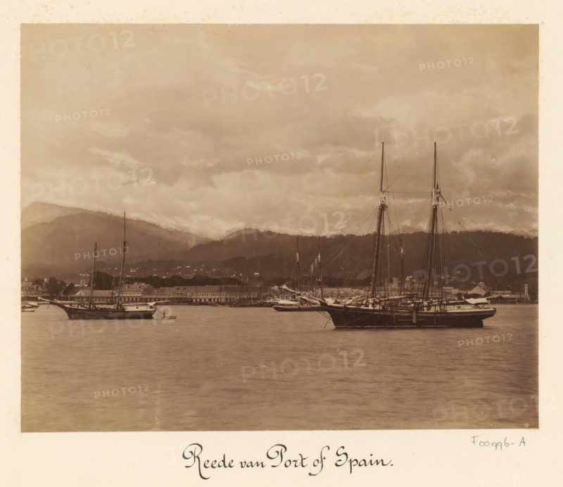 View of the harbor of Port of Spain, album page, photograph, This photo is part of an album., inscription: 'Reede van Port of Spain', Trinidad (Trinidad and Tobago), 1880 - 1900, photographer, paper, albumen print, height 347 mm x width 297 mm, city-view in general, 'veduta', harbor (view, vehicle, ship, etc.), A sepia-toned photograph showing a calm harbor scene with sailing ships moored on the water, tall masts and rigging rising against a cloudy sky, a long waterfront of low buildings along the shore, hills in the background partially covered by clouds, and reflections of the vessels and sky visible on the water surface.