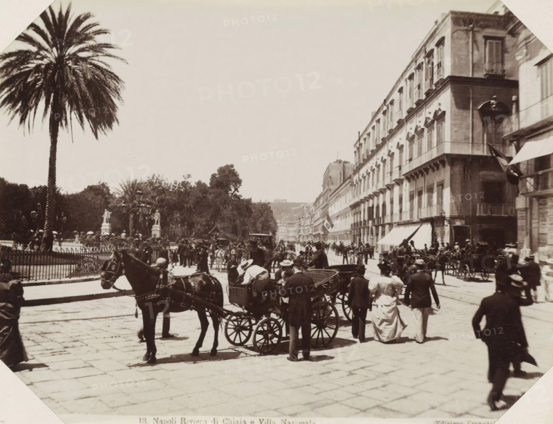 Street Naples, Napoli Riviera di Chiaja e Villa Nazionale, Street in Naples with horse-drawn carriages and passers-by, photograph, Part of Album with 191 photos of a trip to and through South Africa., negative number: '13', Naples, publisher: Edizione Esposito, c. 1880 - c. 1920, photographer, paper, albumen print, albumendruk, height 198 mm x width 258 mm, four-wheeled, animal-drawn vehicle, eg: cab, carriage, coach, street (city(-scape) with figures, staffage), A wide street scene with a large palm tree and fenced garden area on the left and a long row of multi-story buildings with balconies and awnings on the right, crowded with people, horse-drawn carriages, and horses filling the foreground and midground; pedestrians in period clothing walk and stand along the street, some near the carriages, while lamp posts and statues are visible within the garden area and trees rise behind the fencing; the paved street extends into the distance where more buildings and small distant figures ar...