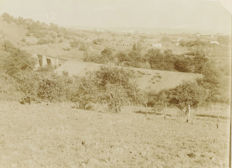 Pretoria, Landscape with railway line near Pretoria, photograph, Part of Album with 191 photos of a trip to and through South Africa., c. 1890 - c. 1920, photographer, paper, daylight gelatin silver print, height 213 mm x width 293 mm, railway, train, A wide pale landscape with rolling hills and scattered trees dominates the scene, with a curving dirt track leading to a low stone bridge spanning a shallow ravine; a few small buildings and structures appear in the distant midground beyond the bridge, and clusters of trees and shrubs fill the slopes and valley below; in the foreground a lone seated man in a hat sits near a bush on grassy terrain dotted with small rocks, and a simple wooden fence with posts runs along the lower right side.