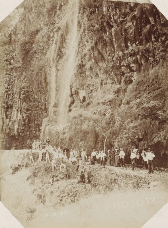 Madeira, Tourist group at a waterfall on Madeira, photograph, Part of Album with 191 photos of a trip to and through South Africa., inscription: 'Rabacal ...', c. 1885 - c. 1910, photographer, paper, albumen print, height 236 mm x width 176 mm, waterfall, A sepia-toned photograph showing a steep rocky cliff covered in grasses and vegetation with a narrow waterfall cascading down its face, and a group of people gathered on a stone path and ledges at the base of the cliff, some standing and some seated, wearing period clothing and hats, with walking sticks and a few small bags, set against the textured rock and plant-covered slope.
