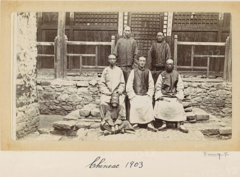 Chinese men and a boy in front of a house in Tibet, photograph, Part of Photo album with 24 photos of army telegrapher DT Dalton's journey through Tibet., title: 'Chinese 1903', photographer: DT Dalton, Tibet, 1903, DT Dalton, photographer, paper, daylight gelatin silver print, height 80 mm x width 137 mm, shoes, sandals, head-gear, child, hairdress, front steps, clothing for the upper part of the body, clothing for the lower part of the body, A sepia photograph shows a group of people posed on uneven stone steps in front of a wooden building with a lattice door and railings; two adults stand on the top step behind three seated adults on a lower step, and a child sits cross-legged at the front smiling; the adults wear long traditional garments with layered tops and wide aprons or robes, and some wear round caps while one holds a small rectangular object; the stone wall and steps have rough, irregular stones and the wooden facade behind them has vertical posts and horizontal beams with ...
