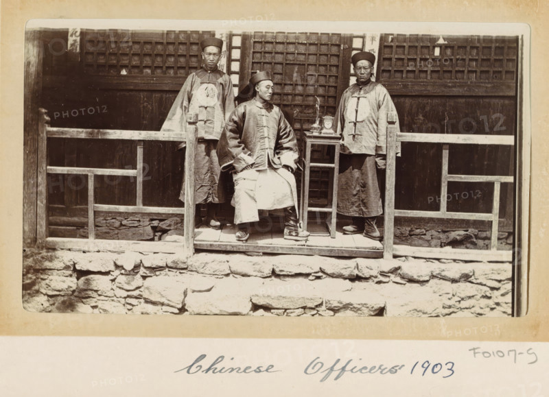 Chinese officers in front of a house in Tibet, photograph, Part of Photo album with 24 photos of army telegrapher DT Dalton's journey through Tibet., title: 'Chinese Officers 1903', photographer: DT Dalton, Tibet, 1903, DT Dalton, photographer, paper, daylight gelatin silver print, height 80 mm x width 137 mm, shoes, sandals, official, civil servant, functional (in general), head-gear, clock, clothing for the upper part of the body, clothing for the lower part of the body, A sepia-toned photograph showing three men wearing traditional long robes and round hats posed on a raised wooden platform in front of a building with wooden lattice windows, a simple wooden railing in front of them, a small table holding a few objects beside the seated man, stonework forming the base of the platform, and the men displaying neutral facial expressions.