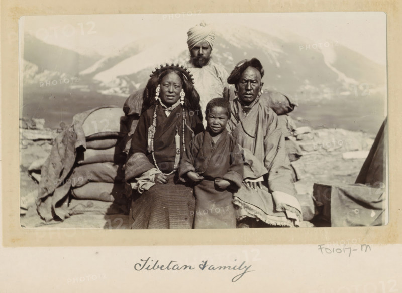 Army intendent with three members of a Tibetan family, Tibet, photograph, Part of Photo album with 24 photos of the journey of army telegraphist DT Dalton through Tibet., title: 'Tibetan Family', photographer: DT Dalton, 1903 - 1906, paper, daylight gelatin silver print, height 80 mm x width 137 mm, head-gear, family, relationship, descent, mountains, clothes covering the entire body, clothing for the upper part of the body, clothing for the lower part of the body, A black and white photograph of four people seated outdoors in front of stacked cloth bundles and a mountainous landscape, with a man wearing a turban standing behind them; the seated adult woman has long hair adorned with a beaded headdress and large round earrings and wears layered garments with long necklaces, the seated adult man wears a fur-trimmed hat and layered robes with a necklace, and a child sits between them wearing a simple robe; the foreground shows fabric textures and folds, the background shows distant snow-...