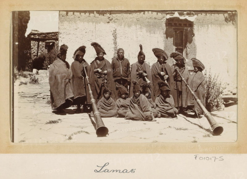 Group of Tibetan lamas with wind instruments (dunchen), Tibet, photograph, Part of Photo album with 24 photos of the journey of army telegraphist DT Dalton through Tibet., title: 'Lamas', photographer: DT Dalton, 1903 - 1906, paper, daylight gelatin silver print, height 80 mm x width 137 mm, head-gear, wind instruments, clothes covering the entire body, priest, non-Christian religions, A group of people arranged in two rows in front of a worn plaster wall, most wearing long cloaks and distinctive tall hats with fur or pointed tops, several seated on the ground wrapped in cloaks, some standing behind them holding long, ornate horns or ceremonial instruments that extend toward the camera, a narrow doorway or window with a decorative frame visible on the right side of the wall, rough stone paving underfoot and an opening to a darker stone structure at the left edge of the scene.