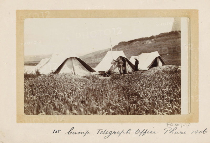 Tent camp of the first British telegraph post in Phari, Tibet, photograph, Part of Photo album with 24 photos of the journey of army telegraphist DT Dalton through Tibet., title: '1st Camp Telegraph Office Phari 1906', photographer: DT Dalton, Phari, 1906, paper, daylight gelatin silver print, height 80 mm x height 137 mm, (military) camp with tents, telegraph pole, telephone pole, (morse) telegraph, A sepia photograph of a row of canvas tents pitched on a grassy plain with taller grass in the foreground and a low hill in the background. Two men stand near one tent, one holding a long pole and the other wearing a hat, and another person lies or sits on the ground nearby. The tents vary in shape with rounded and peaked entrances, and some flaps are open revealing darker interiors. The image is mounted on a light colored card with a rectangular border around the photograph.