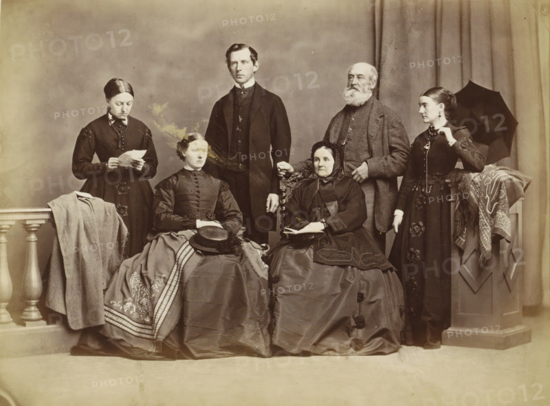 Group portrait of a family in a photo studio, possibly in Norway, photograph, Part of an English family album with photos of people, travels, cricket and artworks., inscription: '...famil...', Norway, c. 1860 - c. 1870, photographer, paper, albumen print, albumendruk, height 156 mm x width 205 mm, historical persons, family group, especially parents with their child(ren), head-gear, beard, dress, gown, skirt, clothing for the upper part of the body, styles of hairdress - AA -, women, umbrella, necklace, studio requisites, clothing for the lower part of the body, A sepia-toned portrait of six people arranged in two rows against a plain backdrop and draped curtain, with three seated women in full skirts and three standing adults behind them; the seated woman on the left wears a dark dress with decorative trim and a patterned shawl draped over her lap with a hat placed on top, the seated woman in the center wears a dark dress with a fitted bodice and has her hands folded, and the seated w...