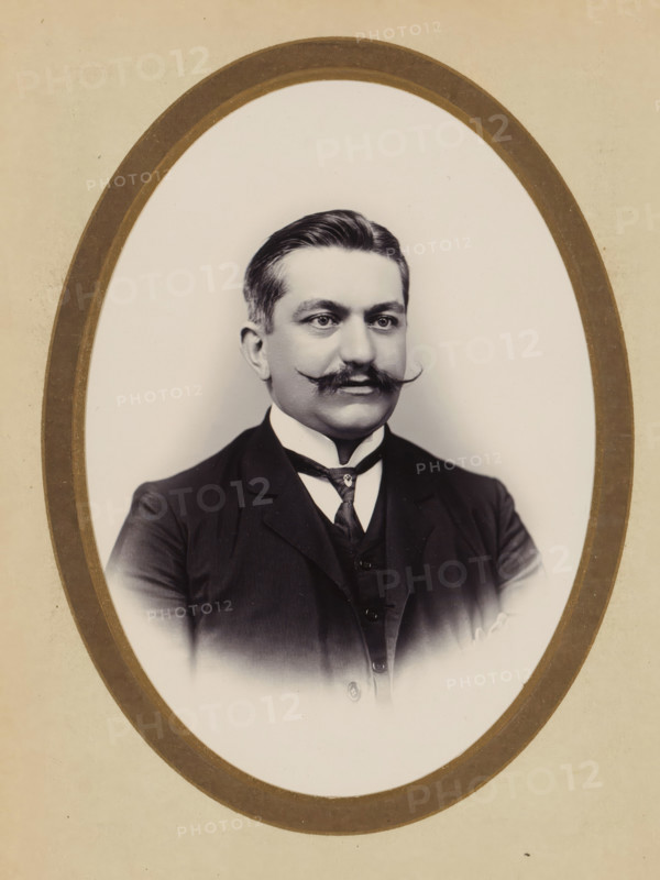 Portrait of a man, album cover, photograph, cabinet photograph, This photo is part of an album., inscription: 'Yu Chong, PEINTRE SUR IVOIRE ET AGRANDISSEMENT, DES PHOTOGRAPHES, 79 RUE DES PANIERS 79, HANOÏ', annotation: 'Lori..e', photographer: Yu Chong, Hanoi, 1880 - 1940, paper, pasteboard, daylight gelatin silver print, height 135 mm x width 96 mm, historical persons, A sepia-toned oval portrait of a man shown from the chest upward, framed by a double oval border; he has neatly combed dark hair, a prominent curled mustache, and a clean-shaven lower face, with light skin and a neutral expression, wearing a dark three-piece suit with a high white collar and a patterned tie, set against a plain light background.