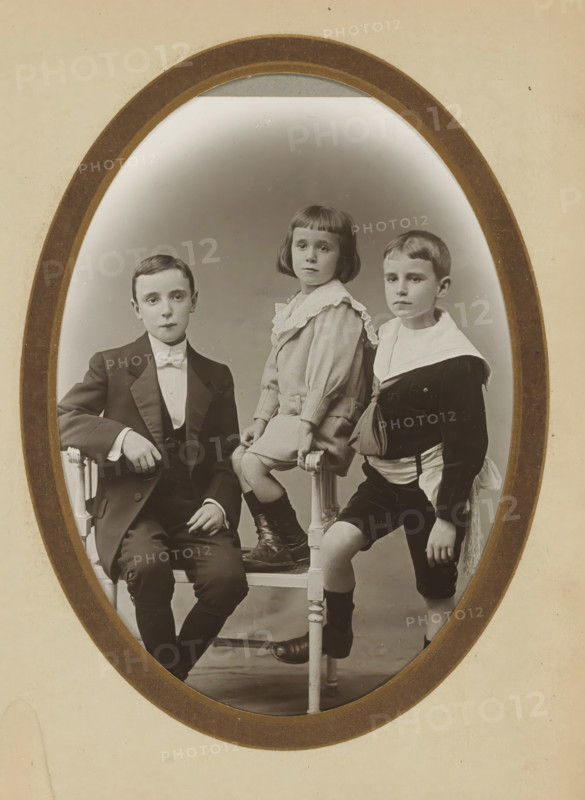 Group portrait of two boys and a young girl, album page, photograph, cabinet photograph, This photo is part of an album., inscription: 'A. Ligey, 21, Rue d'Auteuil, Paris', photographer: Albin Ligey, Paris, 1895 - 1920, Albin Ligey, photographer, paper, pasteboard, matte daylight collodion silver print, height 135 mm x width 96 mm, boy (child between toddler and youth), girl (child between toddler and youth), anonymous historical persons portrayed in a group, in a group-portrait, An oval framed sepia photograph showing three children posed on and around a wooden chair; on the left a boy in a dark suit and bow tie sits with one arm resting on the chair back and a neutral expression, in the center a girl with a bob haircut and a light dress with a ruffled collar sits on the chair seat with her hands resting on the rails and wearing dark boots, and on the right a boy in a dark sailor-style outfit with a light collar stands with one foot on the chair rung and a neutral expression.