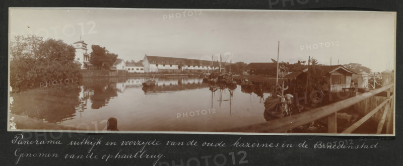 Panorama of the lookout and front of the old barracks in the Lower Town. Taken from the drawbridge., Panorama of the old barracks in the Lower Town of Batavia, seen from the drawbridge, album page, photograph, Part of Travel Album with photos of Batavia., Batavia, c. 1920, photographer, Java, West Java, paper, daylight gelatin silver print, height 248 mm x width 305 mm, barracks, prospect of city, town panorama, silhouette of city, overseas history, colonial history, Dutch East Indies, The, A wide sepia-toned riverside scene showing calm water reflecting buildings and boats, with a row of low whitewashed structures and a long gabled building along the far bank, a taller multi-story building with a tower and surrounding trees on the left, clustered small boats and masts moored near wooden sheds and piers on the right, a wooden railing and walkway in the foreground leading toward a small hut where a person is standing, and vegetation along the river edges under a pale sky.