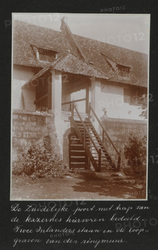 The southern gate with stairs of the barracks mentioned above. Two natives are standing in the trenches of the ring wall., Southern gate with stairs of the old barracks in Batavia, two Javanese are standing in the trenches of the ring wall, album page, photograph, Part of Travel Album with photos of Batavia., Batavia, c. 1920, photographer, Java, West Java, paper, daylight gelatin silver print, height 248 mm x width 305 mm, barracks, gate, entrance, overseas history, colonial history, Dutch East Indies, The, A sepia photograph of a two-story building with a tiled pitched roof and two small dormer openings, showing a covered porch supported by square columns and accessed by a wooden staircase with handrails leading down to an arched doorway set in a stone retaining wall; vegetation grows at the base of the wall and along the foreground, and a person is standing on the porch near the top of the stairs.