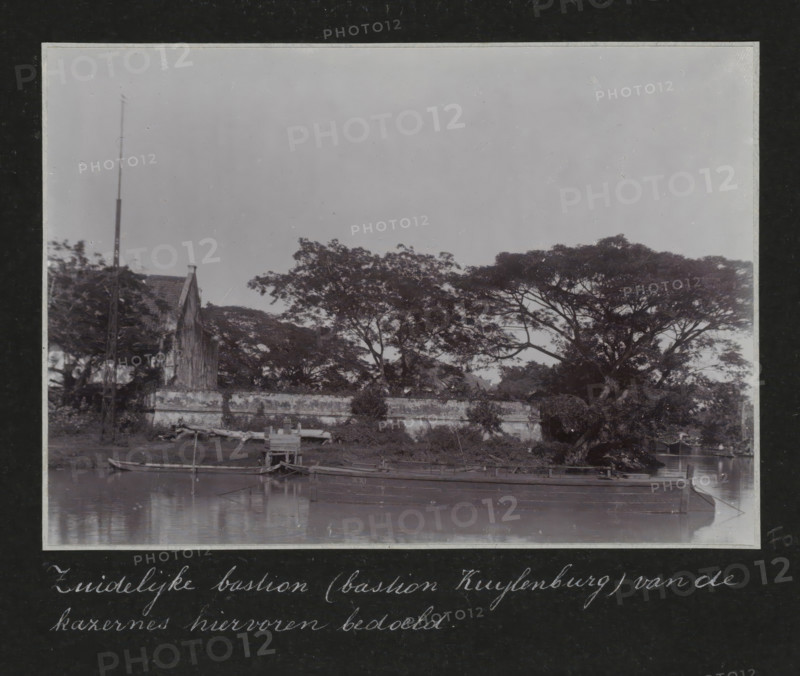 Southern bastion (Bastion Kuylenburg) of the barracks mentioned above., Southern Bastion Kuylenburg at the old barracks in Batavia, album page, photograph, Part of Travel Album with photos of Batavia., Batavia, c. 1920, photographer, Java, West Java, paper, daylight gelatin silver print, height 248 mm x width 305 mm, barracks, overseas history, colonial history, Dutch East Indies, The, A black and white photograph shows a waterfront scene with a calm body of water in the foreground and a low stone or concrete wall running parallel to the shore. Small wooden boats are moored at the water's edge, some tied to simple wooden structures and a short pier. Behind the wall, a cluster of large, spreading trees with dense canopies occupies much of the middle ground, their branches extending horizontally. To the left, a pitched-roof building with a visible gable and a tall, slender pole or mast stands partially obscured by foliage. The sky above is plain and light in tone.