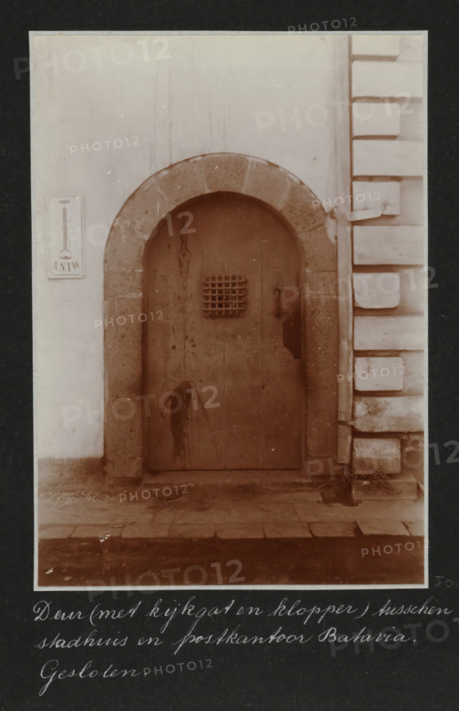 Closed door between the city hall and the post office in Batavia, on the left a sign of the ANIW, Door (with peephole and knocker) between city hall and post office Batavia. Closed., album page, photograph, Part of Travel album with photos of Batavia., Batavia, c. 1920, photographer, Java, West-Java, paper, daylight gelatin silver print, height 248 mm x width 305 mm, gate, entrance, overseas history, colonial history, Dutch East Indies, The, A weathered arched wooden door set into a stone surround within a pale wall, the door showing stains and a small barred opening near its upper center and a metal handle to one side, flanked on the right by a section of wall with deep rectangular block-like projections and on the left by a smooth plastered surface with a small rectangular plaque mounted on it, all seen above a worn stone threshold and paving.
