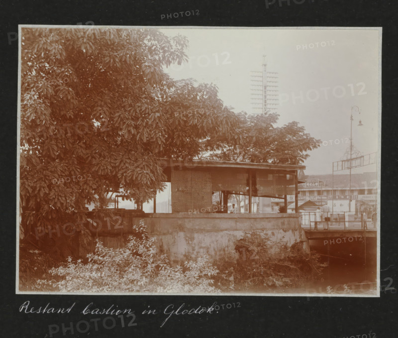 Remnant of a bastion in Glodok., Remnants of a bastion in the Chinese quarter of Glodok in Batavia, in the background modern traffic and advertising for a photo shop, album page, photograph, Part of a travel album with photos of Batavia., Batavia, c. 1920, photographer, Java, West Java, paper, daylight gelatin silver print, height 248 mm x width 305 mm, ruin of a dwelling, house, castle, studio, overseas history, colonial history, Dutch East Indies, The, A sepia-toned scene showing a large leafy tree occupying the left and center foreground with dense foliage, a low concrete wall in front of an open-sided wooden shelter with a corrugated roof and woven panels, a person partially visible sitting inside the shelter, vegetation and low plants growing along the base of the wall, a narrow waterway or dark channel at the right edge beneath a small bridge, a tall vertical pole with multiple horizontal crossarms rising behind the trees, a decorative street lamp on a slender post near the right...
