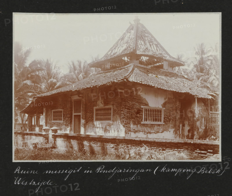 Remains of a mosque (west side) in Pendjaringang (kampong Bèbèk), Ruin of a mosque in Pendjaringang (kampong Bebek) West side., album page, photograph, Part of Travel album with photos of Batavia., Batavia, c. 1920, photographer, Java, West Java, paper, daylight gelatin silver print, height 248 mm x width 305 mm, ruin of a dwelling, house, castle, overseas history, colonial history, Dutch East Indies, The, A weathered brick building with a tiled, multi-tiered roof showing broken and missing tiles and exposed rafters, windows with vertical wooden bars, a narrow doorway, peeling plaster on the exterior walls, a low brick and stucco perimeter wall in front, and tall palm trees visible behind and beside the structure under a pale sky.
