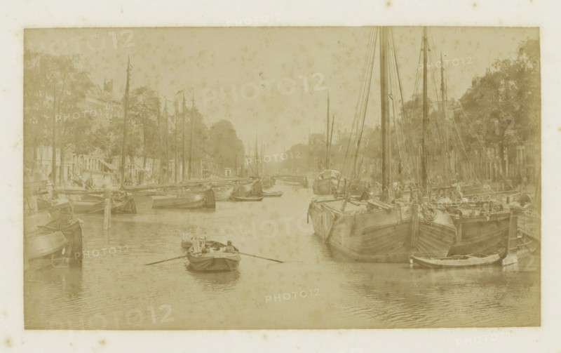 View of the Nieuwehaven in Rotterdam, photograph, Part of Photo album with cityscapes of Rotterdam., inscription: 'Nieuwehaven', Rotterdam, c. 1880 - c. 1900, photographer, paper, albumen print, height 108 mm x width 186 mm, rowing-boat, canoe, sailing-ship, sailing-boat, harbour (view, vehicle, ship, etc.), A sepia-toned scene of a narrow waterway lined with moored sailing vessels on both sides, showing tall masts and rigging, hulls with visible planks and ropes, and smaller rowboats on the water including one with a person rowing toward the foreground; tree-lined quays and dockside buildings are visible behind the boats, and the water surface reflects the vessels and ripples gently.