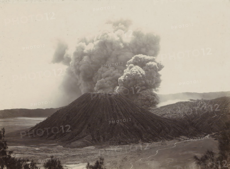 Smoking volcano in the Dutch East Indies, photograph, Part of Travel Album with Forty Photos of the Dutch East Indies., photographer: Onnes Kurkdjian, Dutch East Indies, The, 1912, paper, daylight gelatin silver print, height 175 mm x width 238 mm, volcano, A large conical mountain with steep, deeply ridged slopes sits in the foreground, its summit appearing flat and dark; behind and rising from its base is a massive dense plume of billowing smoke and ash that fills much of the sky with rounded, turbulent shapes; surrounding terrain shows eroded ridges and valleys with a wide flat plain in front, a winding pale stream crossing the plain, and distant low hills along the horizon; the overall palette is muted and monochromatic with textured surfaces and a hazy atmosphere.