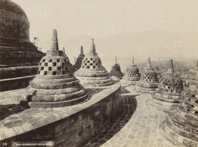 Stupas of Borobudur on Java, photograph, Part of Travel Album with Forty Photos of the Dutch East Indies., number: '25', photographer: Onnes Kurkdjian, Borobudur, 1912, Java, Central Java, paper, daylight gelatin silver print, height 178 mm x width 236 mm, temple, shrine, Hinduism, Buddhism, Jainism, Stone bell-shaped stupas with lattice openings sit atop tiered circular bases arranged along curved stone walkways, their pointed pinnacles rising above carved moldings and layered platforms. A large domed structure with carved bands and weathered blocks occupies the left side, and a person wearing light clothing and a broad-brimmed hat stands on its steps. The stone surfaces show varied tones and weathering, and distant vegetation and faint hills appear on the horizon under a pale sky.