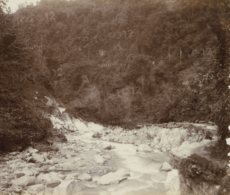 Stream with a road alongside it and workers in the Dutch East Indies, album page, photograph, Part of Album with photos of railways in the Dutch East Indies and postcards., Dutch East Indies, The, c. 1880 - c. 1910, photographer, paper, pasteboard, albumen print, height 230 mm x width 271 mm, railway, train, brook, A rocky riverbed with flowing water running toward the foreground, surrounded by scattered boulders and stones, flanked by steep, exposed rock faces and dense vegetation; the midground shows a mix of shrubs and trees along the banks and a wooden plank or beam lying across rocks, while the background is dominated by a densely forested hillside with tall trees and a few bare branches visible against the canopy.