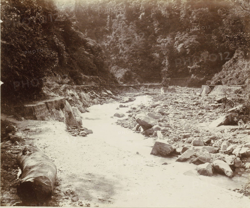 Stream bed with a road in the Dutch East Indies, album page, photograph, Part of Album with photos of railways in the Dutch East Indies and postcards., Dutch East Indies, The, c. 1880 - c. 1910, photographer, pasteboard, paper, albumen print, height 277 mm x width 232 mm, railway, train, brook, A wide rocky riverbed flanked by steep forested hills, with scattered boulders and stones along the shore and in the shallow water, a narrow path or ledge running along the left side beside exposed rock faces, a large fallen log resting near the foreground on the left, dense trees and foliage covering the slopes in the background, and a small man-made structure near the mid-right bank beside the rocky embankment.