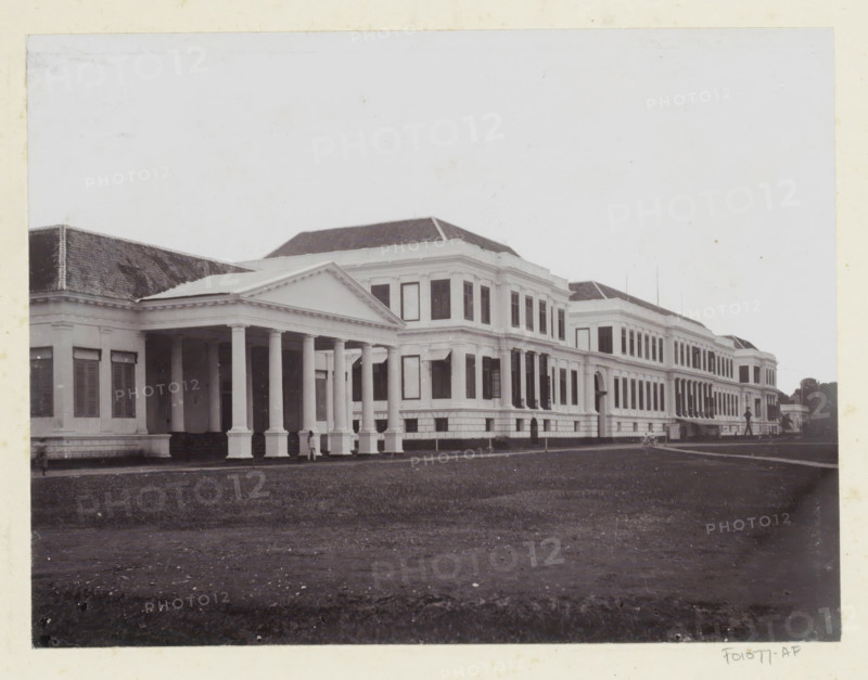 Waterlooplein Palace, Exterior of Daendels' palace on Waterlooplein in Batavia, album page, photograph, Part of Album with photos of Batavia and of the Ursuline sisters., Batavia, c. 1890 - c. 1910, photographer, Java, West Java, paper, pasteboard, daylight collodion silver print, height 170 mm x width 224 mm, palace, A large classical white building stretches across the image with a colonnaded portico at the left featuring tall round columns and a triangular pediment, adjoining a multi-story wing with rows of tall rectangular windows and decorative cornices; the building has a tiled hipped roof and several projecting bays, and stands behind a broad expanse of lawn in the foreground, with a few small indistinct figures near the portico and along the façade.