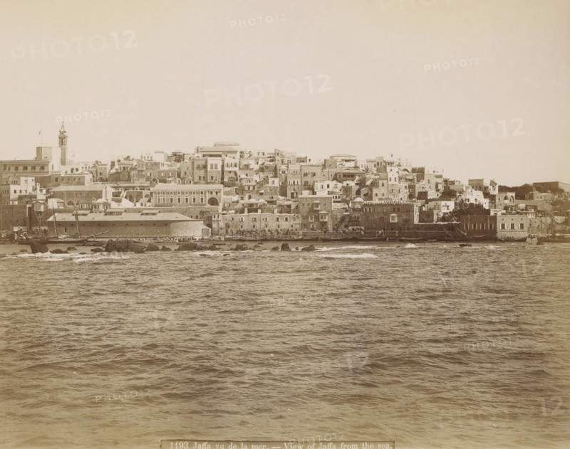 Jaffa, seen from the sea, Jaffa vu de la mer. - View of Jaffa from the sea., photograph, Part of Travel Album with Photos of Jerusalem and Other Biblical Places., negative number: '1193', photographer: attributed to Maison Bonfils, c. 1867 - c. 1895, Ottoman Empire, Attributed to, pasteboard, paper, albumen print, height 222 mm x width 282 mm, prospect of city, town panorama, silhouette of city, A wide waterfront view showing a dense cluster of multi-level stone and plaster buildings along a shoreline, with flat roofs, domes, arched windows and doorways, and a tall tower with a pointed top rising above the other structures, all rendered in a sepia tone; in front of the buildings a narrow quay with small boats and masts is visible, dark rocks protrude at the waterline, and the foreground is filled with rippling sea water under a pale sky.