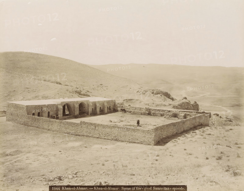 Khan al-Ahmar.- Khan al-Ahmar: Scene of the 'Good Samaritan' episode., Walled building in Khan al-Ahmar, photograph, Part of travel album with photos of Jerusalem and other biblical places., negative number: '1044', photographer: attributed to Maison Bonfils, Khan Al-Ahmar, c. 1867 - c. 1895, Palestine, West Bank, Attributed to, pasteboard, paper, albumen print, height 225 mm x width 283 mm, fence, wall, paling, desert, A stone enclosure sits in a barren landscape, its perimeter formed by low rough stone walls surrounding a rectangular courtyard and a long building with a flat roof and a series of arched openings along one side; inside the courtyard and near the arches are a few animals and figures, and a person stands near the outer wall; the surrounding terrain is dry and rolling with sparse vegetation, rocky outcrops, and distant undulating hills under a pale sky.