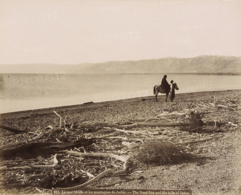 Rider and man on the shore of the Dead Sea with washed-up branches in the foreground, La mer Morte et les montagnes de Judée.- The Dead Sea and the hills of Judea., photograph, Part of Travel album with photos of Jerusalem and other biblical places., negative number: '911', photographer: Maison Bonfils, Dead Sea, c. 1867 - c. 1895, Jordan, pasteboard, paper, albumen print, height 227 mm x width 284 mm, beach, sea (landscape with figures, staffage), A wide shoreline scene with a pebbled beach strewn with pieces of driftwood in the foreground, a calm body of water extending to a distant mountain range under a pale sky, and a person mounted on a horse near the water's edge accompanied by another standing figure beside the horse.