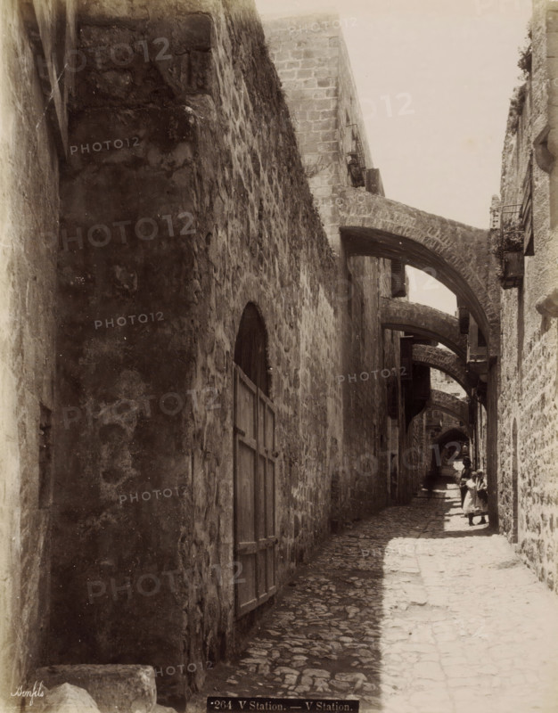 Fifth Station of the Cross on the Via Dolorosa in Jerusalem, V Station. - V Station., photograph, Part of Travel Album with Photos of Jerusalem and Other Biblical Places., negative number: '264', photographer: Maison Bonfils, Jerusalem, c. 1867 - c. 1895, pasteboard, paper, albumen print, height 282 mm x width 223 mm, street, stations of the cross, Via Dolorosa, A narrow cobblestone alley flanked by tall stone walls with multiple stone arches spanning overhead, wooden shutters and doors set into the walls, sunlight casting a bright strip along the center of the path while the sides remain in shadow, and several small figures farther down the alley including one adult and children wearing simple garments standing in the sunlit area.