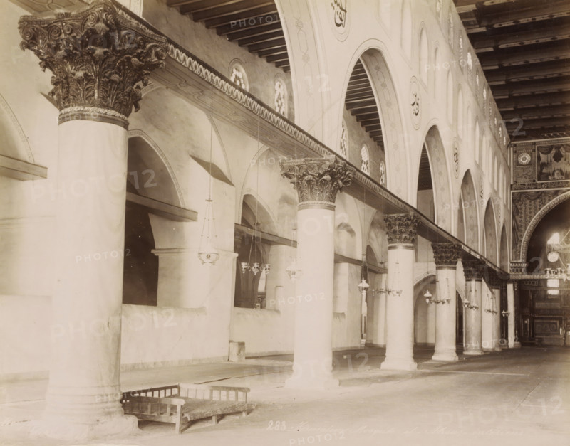 Jerusalem. Mosquée el-Aksa, - intérieur, Interior of the Al-Aqsa Mosque on the Temple Mount in Jerusalem, photograph, Part of Travel album with photos of Jerusalem and other biblical places., inscription: 'BFK Rives', negative number: '283', photographer: Maison Bonfils, Jerusalem, c. 1867 - c. 1895, pasteboard, paper, albumen print, height 225 mm x width 282 mm, temple, shrine, Islam, Mohammedanism, Al-Aqsa Mosque, A long interior view showing a row of tall cylindrical columns with ornate carved capitals supporting a decorated horizontal beam and a high ceiling with exposed wooden beams, a series of arched openings along the wall each topped by small circular windows, hanging pendant lights suspended from the ceiling at various heights, decorative painted or carved roundels on the arches, patterned detailing along the beam and arch faces, a wooden bench resting near the base of one column, and a richly decorated archway and panel work visible toward the far end.