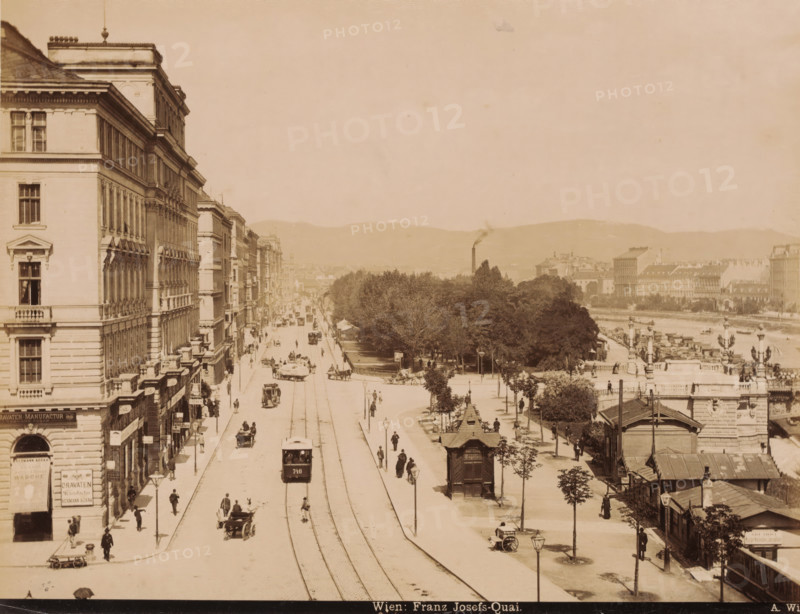 View of the Franz Josefs-Quai. in Vienna with shops, buildings, trams and carts, Wien. Franz Josefs-Quai., photograph, Part of Travel album with photos of sights in Germany and Austria and of works of art., photographer: A. Wimmer, Vienna, c. 1880 - c. 1895, Lower Austria, paper, albumen print, height 202 mm x width 264 mm, shop, store, tramway, tram, pier, quay, wharf, four-wheeled, animal-drawn vehicle, eg: cab, carriage, coach, street (city(-scape) with figures, staffage), A wide street scene showing a long avenue flanked by tall multi-story buildings on the left and a tree-lined promenade and riverbank on the right, with tram tracks running down the center of the street and a tram car moving along them; horse-drawn carriages and pedestrians are visible on the road and sidewalks, lamp posts line the avenue, and small kiosks and structures dot the promenade; in the distance low hills form the horizon and a smokestack rises above clustered buildings, while decorative facades, balconie...