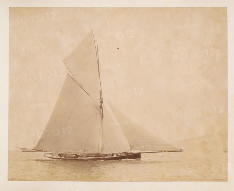 Sailing ship launched, album page, photograph, This photo is part of an album., check stamp: 'Adamson, Rothesay.H.B', photographer: James Adamson, c. 1880 - c. 1900, paper, albumen print, height 215 mm x width 272 mm, sailing-ship, sailing-boat, landscapes with waters, waterscapes, seascapes (in the temperate zone), A large sailboat shown in profile on calm water with tall sails raised and visible rigging, a long mast rising toward the top of the image, a sleek dark hull with details of the deck and small structures, thin lines of the sails showing seams and shading, a small boat alongside the main vessel, a faint distant shoreline on the left, and a pale, textured sky occupying much of the background.
