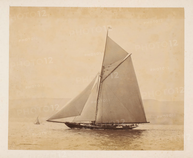 Two sailing ships launched, album page, photograph, This photo is part of an album., check stamp: 'Adamson, Rothesay.H.B', photographer: James Adamson, c. 1880 - c. 1900, paper, albumen print, height 229 mm x width 279 mm, sailing-ship, sailing-boat, landscapes with waters, waterscapes, seascapes (in the temperate zone), A large sailboat viewed in profile on calm water with its tall mast and multiple taut sails filling the right side of the image, a small flag at the mast top, rigging lines and spars visible, crew members positioned along the deck, a smaller distant sailboat on the left horizon, soft tonal gradations in the sky and faint landforms near the horizon, and the photograph framed by a light border with subtle aging marks.