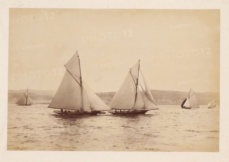 Various sailing ships, including Woodcock and Yvonne, at sea with a hilly landscape in the background, album page, photograph, This photo is part of an album., check stamp: 'Adamson, Rothesay.H.B', photographer: James Adamson, c. 1880 - c. 1900, paper, albumen print, height 191 mm x width 283 mm, sailing-ship, sailing-boat, landscapes with waters, waterscapes, seascapes (in the temperate zone), (high) hill, Several sailboats are shown on calm water, each with tall masts and large triangular sails filled by the wind, two of the boats positioned close together in the foreground with visible hulls and small groups of people sitting along the decks, and additional sailboats farther back near a distant shoreline with low hills; the sky above is clear and light, and the overall image has a sepia tone.
