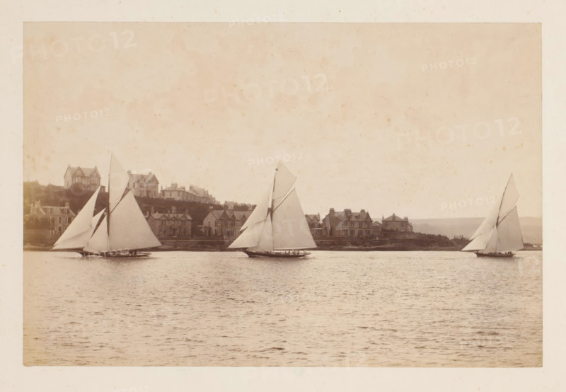 Sailing ships Yvonne, Doris and Darthula in the water with buildings in the background, album page, photograph, This photo is part of an album., check stamp: 'Adamson, Rothesay.H.B', photographer: James Adamson, c. 1880 - c. 1900, paper, albumen print, albumendruk, height 182 mm x width 284 mm, sailing-ship, sailing-boat, landscapes with waters, waterscapes, seascapes (in the temperate zone), civic architecture, edifices, dwellings, A sepia-toned scene of a calm body of water with three sailboats in the foreground, each boat showing tall triangular sails and low hulls with small groups of people aboard; along the distant shoreline a row of houses and larger buildings sit on a gentle rise with varied rooflines and chimneys, and grassy areas and trees are visible behind and between the structures; the sky above is light and mottled with faint spots and a textured appearance.