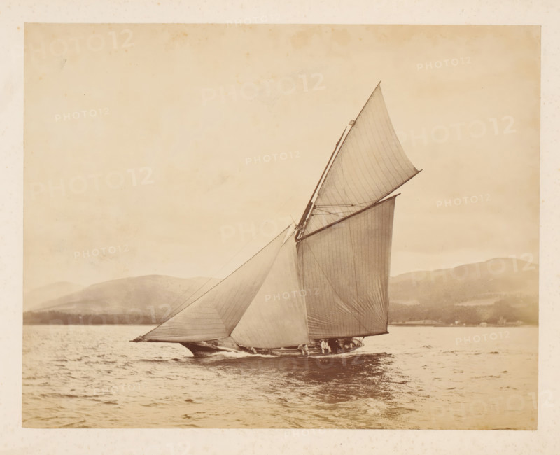 Sailing ship Idalia No. 1 launched with a hilly landscape in the background, album page, photograph, This photo is part of an album., check stamp: 'Adamson, Rothesay.H.B', photographer: attributed to James Adamson, c. 1880 - c. 1900, Attributed to, paper, albumen print, height 218 mm x width 279 mm, sailing-ship, sailing-boat, landscapes with waters, waterscapes, seascapes (in the temperate zone), (high) hill, A large sailboat pictured on a body of water with tall, triangular sails filled by wind, rigging lines visible against a light sky, a low shoreline and hills in the background, rippled water in the foreground, and a narrow strip of boat hull and crew partially visible near the waterline.