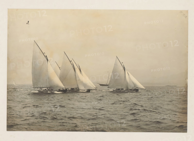 Various sailing ships, including the Windfall, launched, album page, photograph, This photo is part of an album., check stamp: 'Adamson, Rothesay.HB, Copyright', number: '3', photographer: attributed to James Adamson, c. 1880 - c. 1900, Attributed to, paper, gelatin silver print, developed gelatin silver print, height 190 mm x width 281 mm, sailing-ship, sailing-boat, landscapes with waters, waterscapes, seascapes (in the temperate zone), Several sailboats with tall triangular sails are shown on open water, their hulls cutting through small waves and producing spray, the sails mostly light in tone with visible seams and lines, a larger boat appears behind them, the sky above is pale and uniform, and the scene fills a framed photographic border.