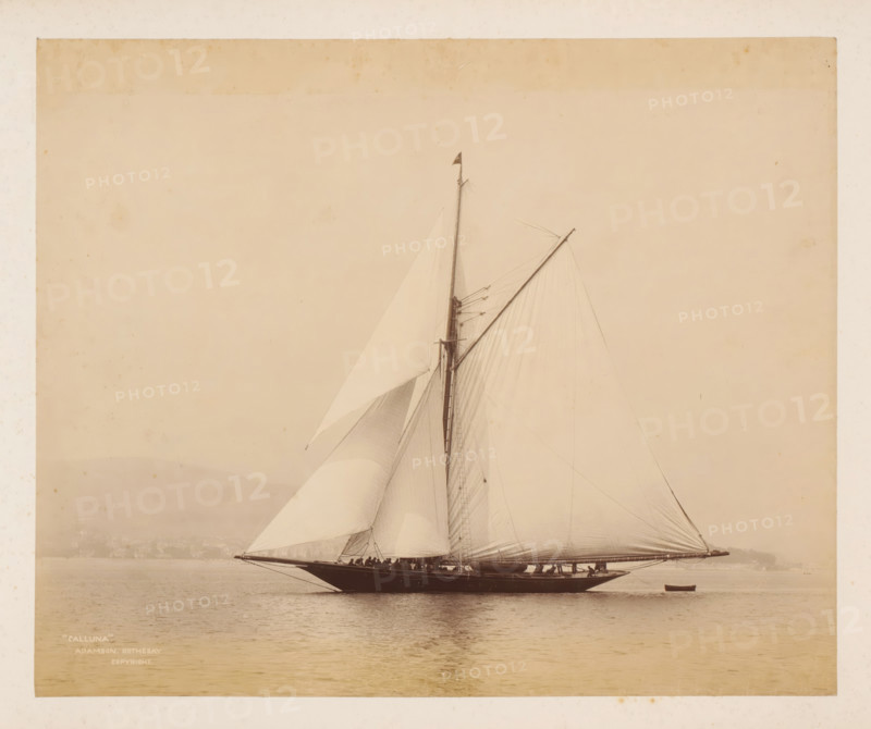 Sailing ship Calluna launched, album page, photograph, This photo is part of an album., inscription: ''Calluna', Adamson. Rothesay., Copyright', photographer: James Adamson, c. 1880 - c. 1900, paper, albumen print, height 229 mm x width 279 mm, sailing-ship, sailing-boat, landscapes with waters, waterscapes, seascapes (in the temperate zone), (high) hill, A large sailboat with tall masts and billowing sails is shown on calm water, its dark hull reflecting faintly on the surface; crew members stand along the deck and rigging lines run from the masts to the bow and stern, while a small rowboat floats nearby and a hazy shoreline and low hills are visible in the background under a pale sky.