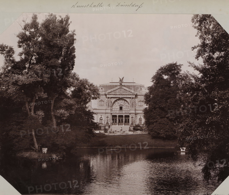 View of the Kunsthalle in Düsseldorf from 1891, album page, photomechanical print, This print is part of an album., annotation: 'Kunsthalle in Düsseldorf', inscription: 'Stengel & Co, 1897, Dresden', Düsseldorf, cliché maker: Stengel & Co., Dresden, photographer, North Rhine-Westphalia, Saxony, paper, collotype, photogravure, height 258 mm x width 355 mm, permanent exhibition, museum, A large neoclassical building with ornate carvings and a central pediment sits beyond a grassy bank, framed by tall leafy trees on both sides and reflected in a calm body of water in the foreground; a stone staircase leads up to the building's entrance where a few small human figures stand near doorways, and an angelic statue crowns the roof above sculptural reliefs, while dense foliage and tree trunks fill the image margins and the water shows rippled reflections of the scene.