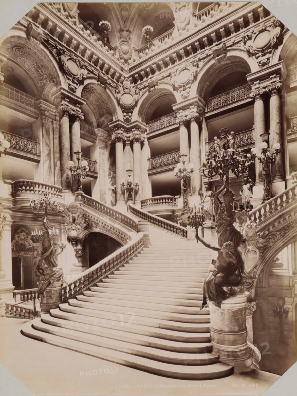 Interior with stairs of the Opéra de Paris, album page, photograph, This photo is part of an album., inscription: '478 - Paris. l'escalier du Grand-Opéra. Ed. H. édit', photographer: Edouard Hautecoer (possibly), Paris, publisher: Edouard Hautecoer, 1890 - 1898, Edouard Hautecoer, photographer, publisher, paper, albumen print, albumendruk, height 258 mm x width 355 mm, staircase, opera house, Opéra de Paris, A wide marble staircase with sweeping curved steps ascends between ornate balustrades, flanked by sculpted figures mounted on pedestals holding candelabras; tall Corinthian columns rise behind the balustrades supporting a richly decorated entablature with carved medallions and reliefs, and a series of large rounded arches reveal upper-level balconies with intricate wrought-iron railings; elaborate chandeliers and multiple lamp fixtures with globes hang and stand throughout the space, while carved ornamental details, garlands, and festoons decorate the arches and vaults above.