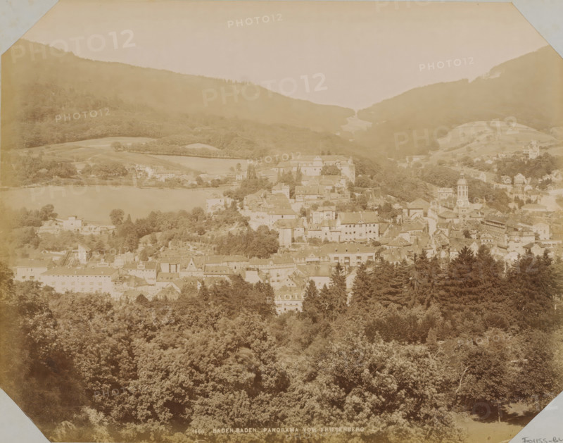 View of Baden-Baden, album page, photograph, This photo is part of an album., annotation: 'Reis Herrenalb. Baden-Baden, Wiesbaden. August 1899', inscription: '1666 Baden-Baden Panorama vom Friesenberg', photographer: LL, Baden-Baden, 1890 - 1899, Baden-Württemberg, paper, albumen print, albumendruk, height 258 mm x width 355 mm, city-view in general, 'veduta', A wide sepia-toned panorama shows a town nestled in a valley with rooftops and clustered buildings rising from a wooded foreground toward a lakeshore and surrounding hills, with a church tower and various multi-story structures among trees, terraced fields and patches of open land on the slopes, dense forest in the foreground and tree-covered hills framing the scene under a pale sky.
