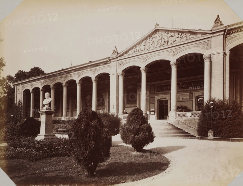 View of the Trinkhalle in Baden-Baden, album page, photograph, This photo is part of an album., inscription: '548 Baden-Baden. - Die Trinkhalle - LL', photographer: LL, Baden-Baden, 1890 - 1899, Baden-Württemberg, paper, albumen print, albumendruk, height 258 mm x width 355 mm, watering-place, spa, A sepia-toned photograph of a classical building facade with a colonnade of tall columns supporting arched openings and an ornate pediment with sculpted reliefs and seated figures, a broad staircase leading up to an entrance flanked by columns, decorative panels and framed artworks visible on the interior walls set back under the arches, manicured shrubbery and small conical trees in the foreground, a stone pedestal topped by a white bust near the left side, a lamppost near the right side, and a pathway curving in front of the stairs.