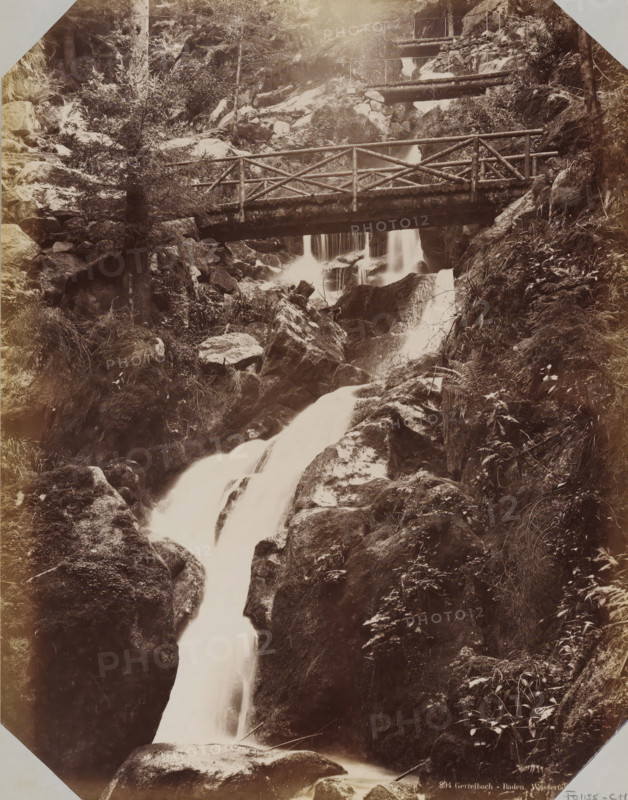 View of the Gertelbach Waterfalls, album page, photograph, This photo is part of an album., inscription: '894 Gertelbach - Baden. Waterfalls. LL', photographer: LL, Baden-Baden, 1890 - 1899, Baden-Württemberg, paper, albumen print, height 258 mm x width 355 mm, waterfall, A sepia-toned photograph showing a rocky forested ravine with cascading water flowing downward over smooth rock surfaces and between boulders, a wooden footbridge with a simple log railing crossing the stream partway up, trees and dense vegetation clinging to the steep rock faces on either side, smaller wooden railings and walkways visible higher up along the slope, wet moss and ferns on the rocks, and streaks of flowing water captured with a silky appearance.