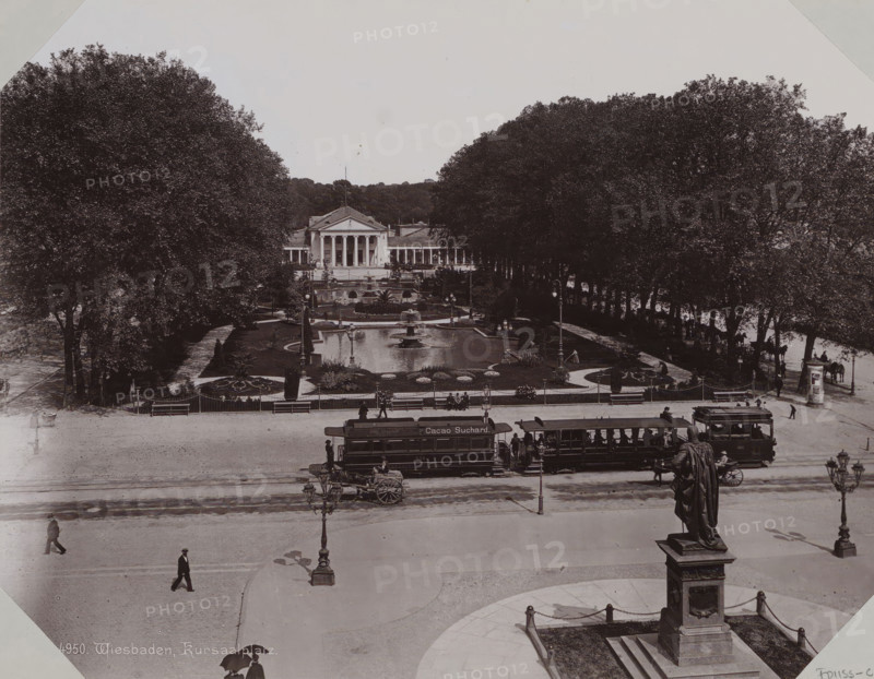 View of the Kurhaus in Wiesbaden, album page, photomechanical print, This print is part of an album., inscription: '4950. Wiesbaden, Kursaalplatz.', 1890 - 1899, paper, collotype, photogravure, height 258 mm x width 355 mm, public gardens, park (city(-scape) with figures, staffage), civic architecture, edifices, dwellings, Hessen, Wiesbaden, A wide plaza scene with a large neoclassical building centered in the background, flanked by rows of tall trees and a formal garden with paths, ornamental flowerbeds, and a rectangular reflecting pool with a central fountain. In front of the garden there are benches, lampposts, and a low decorative fence, while tram cars and a horse-drawn cart are positioned along the street in front of the plaza. Several pedestrians walk across the open paved area, and a large statue on a pedestal stands in the foreground with chained posts around its base. The image is monochrome and shows a broad view from an elevated vantage point.