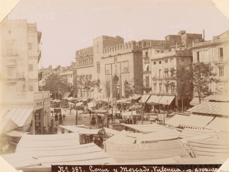 Market in Valencia, Lonja y Mercado. Valencia, album page, photograph, This photo is part of an album., number: '187', annotation: 'Valencia - Marketplace and Bullring', photographer: Antoni Esplugas, Valencia, c. 1888 - in or before 1892-04-07, paper, albumen print, height 161 mm x width 214 mm, market, A wide street scene showing rows of market stalls with canvas canopies extending across the foreground and middle ground, a crowd of people moving between the stalls, a few horse-drawn vehicles among them, and taller buildings lining the background including a large ornate façade with arched doorways and decorative stonework flanked by multi-story residential and commercial buildings with balconies and windows, as well as several trees rising between the buildings.