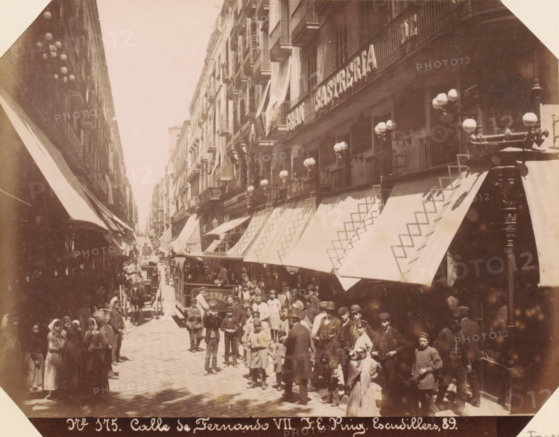 Street view of Calle de Ferran in Barcelona, Calle de Fernando VII, album page, photograph, This photo is part of an album., annotation: 'Barcelona Calle de Fernando', number: '175', photographer: JE Puig, Barcelona (city), c. 1885 - in or before 1892-04-09, Catalonia, paper, albumen print, height 167 mm x width 214 mm, street, A sepia-toned street scene shows a crowded narrow avenue flanked by multi-story buildings with rows of balconies and large fabric awnings extending over the sidewalk; many globe street lamps protrude from the buildings and ornate lampposts line the street; a dense group of people stands and moves along the cobbled road, including adults and children wearing period clothing and hats; a horse-drawn vehicle and a streetcar are visible among the crowd; shopfronts and awnings display geometric patterns and there are draped fabrics hanging from some balconies.