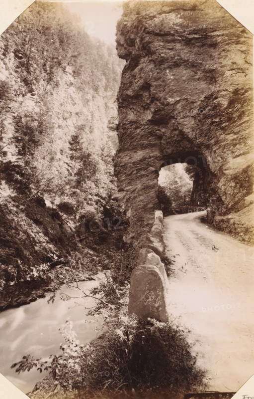 Taminakloof, album page, photograph, This photo is part of an album., annotation: 'Ragatz - Tamina Schlucht', c. 1885 - in or before 1892-04-29, photographer, paper, albumen print, height 209 mm x width 132 mm, ravine, chasm, abyss, canyon, A narrow dirt road curves into a tunnel carved through a tall rocky cliff, with a low stone barrier lining the road's outer edge; the tunnel opening reveals light and trees beyond while the cliff face shows layered rough textures and overhangs; opposite the road a steep forested slope rises with dense trees and undergrowth, and a stream flows at the base past rocks and vegetation; the scene is rendered in a sepia-toned photograph with soft contrasts and a vignette around the edges.