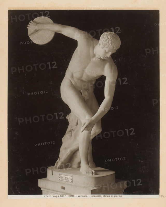 Roma - Vaticano - Discus Thrower, marble statue, Sculpture of a discus thrower in the Vatican Museums in Vatican City, photograph, Part of Photo album with photographs of sights and artworks in Italy., number: '4067', photographer: Edizioni Brogi, Vatican Museums, c. 1875 - in or before 1907, Vatican City, paper, gelatin silver print, height 250 mm x width 198 mm, sculpture, discus-throwing, A light-colored marble statue of a nude male athlete is shown against a dark background, captured in a dynamic throwing pose with one arm extended backward holding a circular disc and the other arm reaching down toward a bent leg, the head turned downward and to the side with stylized curly hair and a focused facial expression, the muscular torso and limbs sculpted with visible anatomical detail, one foot planted on a round base and the other leg slightly raised and supported by a tree-trunk-like element behind the figure, the base rests on a stepped plinth and the image has a narrow light-colored ...